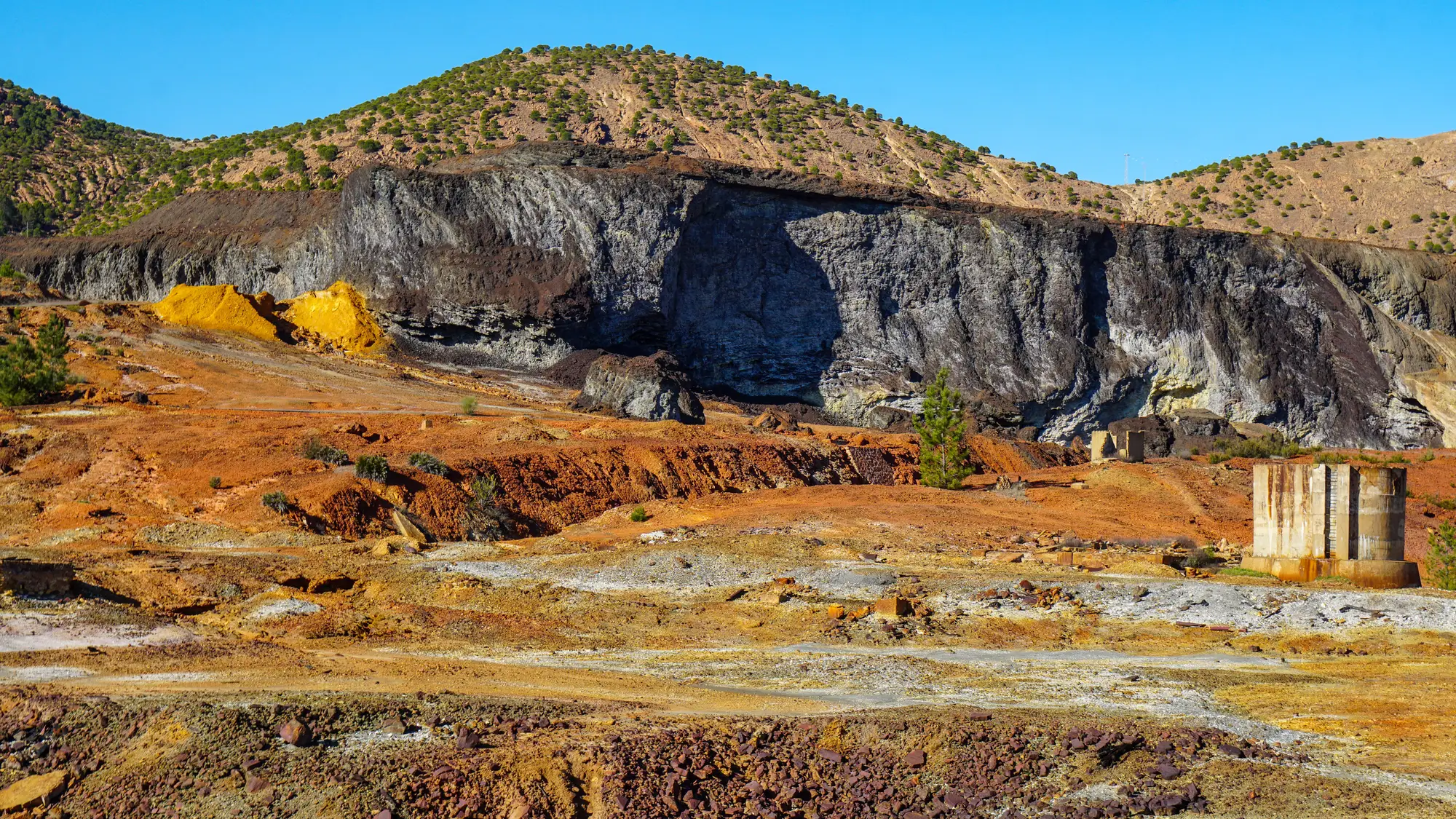 Disused workings on a dry hillside: a grey quarried face, orange and yellow spoil, and the ruin of an old concrete structure